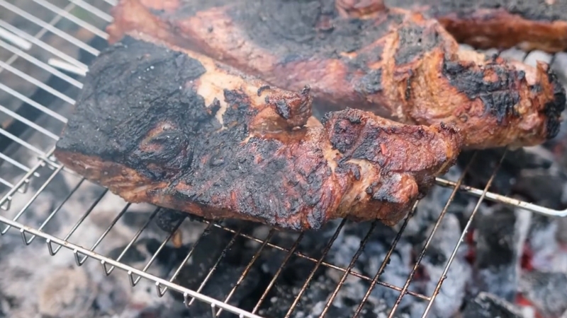 Charred beef ribs rest on a grill over glowing coals, releasing smoke and rich aroma