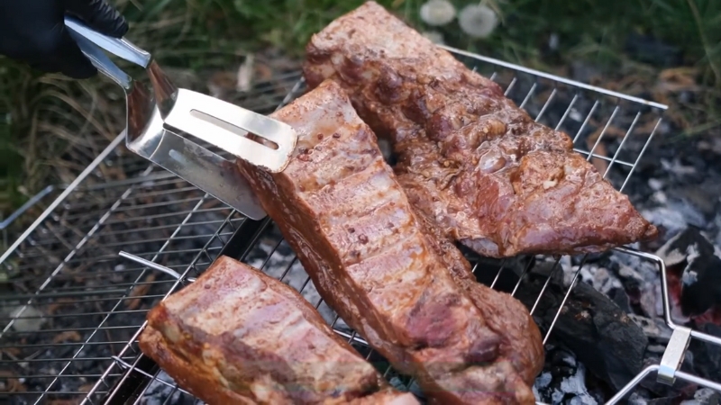 Beef ribs are lifted with tongs over a charcoal grill, showing a smoky sear