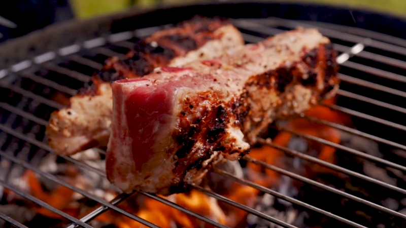 A thick beef rib sears over open flames on a grill, showing a charred crust with a rare center