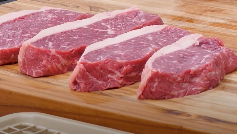Four raw steaks on a wooden cutting board beside a sharp knife